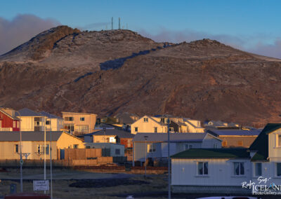 In the image, there is a mountainous landscape with a rugged, rocky terrain. A snowy peak rises in the background, where communication towers can be seen perched at the top. In the foreground, a collection of houses is visible, featuring a mix of architectural styles and colors, including red, white, and green. The houses are arranged closely together, some with visible wooden fences, and they sit against the backdrop of the mountain. The scene is illuminated by warm light, suggesting either early morning or late afternoon. The sky above is clear with subtle blue tones.