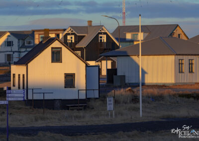 The image shows a row of houses in a small town setting, with a mix of architectural styles and colors. In the foreground, there is a white house with a sloped black roof, partially lit by the sun, creating strong shadows and highlights. To the left, another house of a similar style but with a darker wooden exterior and a balcony is visible. Behind this, more houses of varying colors and designs can be seen, indicating a close-knit community. A signpost is positioned in the foreground, suggesting the presence of nearby roads or paths. The ground is covered with dry grass, and the overall atmosphere is peaceful and slightly rustic, characteristic of a residential area. The sky above is lightly clouded, contributing to a serene evening or early morning ambiance.