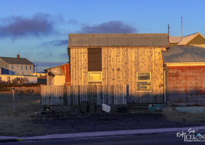 The image depicts a rustic building with a weathered wooden exterior, illuminated by the warm glow of the setting sun. The structure features a metal roof, with sections of the wall painted in a reddish hue. To the left, there is a fenced area with wooden slats, and a few scattered objects, including a white container or cooler. Surrounding the building are patches of dry grass, and in the background, there are additional buildings and a communication tower. The sky above is clear with soft clouds, creating a serene atmosphere.