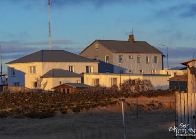 The image shows a cluster of buildings set against a clear sky. Two main houses are prominently featured: one has a white, corrugated exterior and a slightly sloped roof, while the other is a mix of grey and blue with multiple windows. In front, there is a small wooden structure, possibly a shed, next to a stone wall covered in moss. Utility poles and a tall communication tower with red and white markings rise in the background, connected by thin wires stretching across the sky. The foreground features dry grass, indicating a rural setting. The scene is illuminated by warm sunlight, creating distinct shadows.