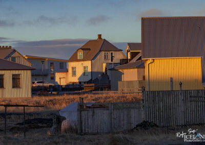 The image depicts a quiet residential scene in a small town, possibly in Iceland. Various houses with different roofing styles and colors are positioned close together. The foreground features a wooden fence, and some areas are bathed in warm sunlight, highlighting the textures of the buildings. The architecture includes a mix of traditional and modern elements, with some houses painted in muted hues and others reflecting a brighter yellow. In the background, there are glimpses of open sky and clouds, suggesting a serene early morning or late afternoon atmosphere. A few cars are parked along the road, and the surrounding landscape is dry with sparse grass, enhancing the tranquil rural setting.