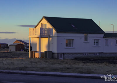 A two-story house stands in a rural setting, with a light-colored exterior and a sloped green roof. The house features a small balcony with wooden railings on the upper level. Sunlight casts a warm glow on the house, highlighting its details against a backdrop of open grassland. In the distance, there are other buildings visible, adding to the serene landscape. The sky is clear, transitioning into twilight, with soft colors blending above.