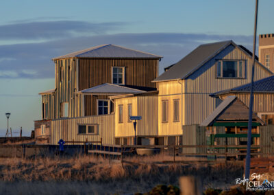 The image showcases a collection of buildings, primarily constructed with metal siding exhibiting a variety of colors, primarily light grey and yellowish hues, illuminated by warm sunlight. The structures have distinct architectural features, including sharp roof angles and several windows. In the foreground, there's a grassy area, and a wooden fence borders the scene, adding to the rural atmosphere. A street sign labeled "Kirkjustræti" is visible, along with a lamp post nearby, indicating proximity to a road. In the background, there are clouds softly dispersed across the sky, creating a calm and scenic backdrop.