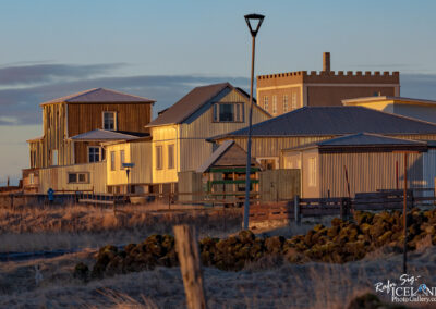 A row of distinct buildings stands against a soft evening sky. The structures, featuring a mix of architectural styles, include a tall, rectangular building with a decorative roof that resembles battlements. The other buildings are predominantly clad in corrugated metal, reflecting warm golden hues from the setting sun. A street lamp nearby casts a gentle glow, while the landscape is framed by sparse grass and rocky terrain, creating a serene, almost rustic atmosphere. The scene conveys a sense of quiet charm and the beauty of simple, functional design in an open, natural setting.