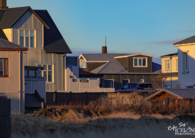 The image showcases a serene, urban landscape featuring several distinctive houses. The architecture is characterized by a mix of traditional and contemporary styles. Some houses have slanted roofs and are clad in either white or muted colors, while others are painted in shades of gray. There are windows reflecting warm sunlight, creating a play of light and shadow on the buildings. In the foreground, patches of dry grass and sparse vegetation contrast with the structured elements of the houses. The background shows a clear blue sky, adding to the calm and inviting atmosphere of the scene.