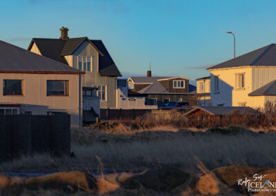 The image shows a quiet residential area with several houses, mostly modern, featuring distinct architectural styles. Some buildings have pitched roofs, while others have flat roofs, and they are painted in shades of white, gray, and green. In the background, there's a clear blue sky with warm sunlight illuminating the structures, creating soft shadows. The foreground is composed of long grass and a rocky surface, adding a natural element to the scene. There are a few glimpses of cars parked in driveways and some sparse vegetation around the houses, contributing to the serene atmosphere.