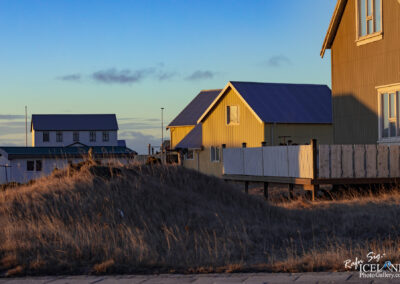The image shows a scenic view of a small, quiet neighborhood at sunset. In the foreground, there is a grassy hillside with golden-brown wild grass swaying gently. On the right, several houses are featured, including a bright yellow house with a sloping roof and a white picket fence. Nearby, a couple of white houses with green roofs stand close together, creating a cozy village atmosphere. The sky above is painted in various shades of blue and soft orange, reflecting the warm light of the setting sun.