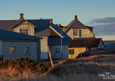 The image features a cluster of houses set against a clear blue sky during what appears to be golden hour, capturing warm light that highlights the buildings. The houses are characterized by textured metal roofs and walls, primarily in shades of blue and gray, creating a rustic yet charming aesthetic. In the foreground, a patch of dry grass and scattered boulders adds to the natural landscape, while a lone streetlamp stands amongst the buildings. The scene evokes a sense of tranquility, typical of a quiet town or village.