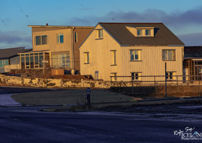 The image shows two distinct buildings alongside a road. The house on the right is a tall, white structure with a peaked roof and multiple windows, reflecting warm sunlight, giving it a cozy appearance. To the left, there's a more modern building with a flat roof, grayish exterior, and large windows that suggest a contemporary design. In front of the buildings, there is a low stone wall, and nearby, a street sign marked "Túngata" stands next to a small grassy area. The road curves gently around the scene, highlighting the peaceful, residential setting.