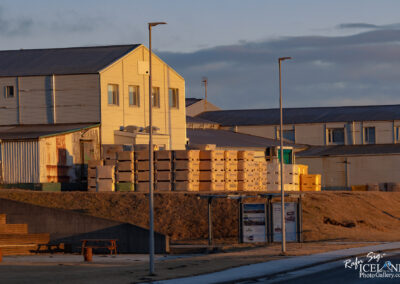 A scene depicting a collection of industrial buildings during sunset. The buildings are primarily made of metal and have a weathered, metallic appearance, featuring various shades of gray and white. On the left, there is a rusted structure with an uneven roof, while in the foreground, several large, stacked boxes are neatly arranged. Two street lamps stand tall, illuminating the area. There’s a small, wooden bench nearby, positioned against a concrete wall, and informational boards about local attractions can be seen, indicating the area is likely a harbor or industrial site. The sky exhibits soft, warm hues from the setting sun, casting a gentle light over the entire scene.