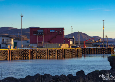 The image depicts a harbor scene during the early evening, capturing a still body of water that reflects the soft colors of the sky. On one side, there are large, round rubber tires stacked along the edge of a dock, providing a buffer for boats. A red building with large windows dominates the background, which looks like it houses some industrial operations, including what appears to be a small set of stairs on one side. Nearby, there are several crates arranged neatly, suggesting some storage activity, along with a vehicle parked on the dock. In the distance, gentle hills rise against the sky, indicating a rugged landscape, and the overall ambiance conveys a serene and industrious harbor atmosphere.
