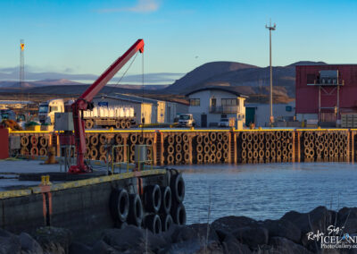 The image depicts a harbor scene at dawn, featuring a yellow dock with a red crane on the left side. Several large tires are stacked along the dock's edge, contributing to the maritime setting. In the background, there are several buildings, including a white structure that appears to be an office, and a red building with a loading area. A truck, likely transporting goods, is parked near the front. The calm water reflects the colors of the sky, which transition from soft blue to hints of orange, suggesting the early morning light. In the distance, rolling hills rise, partially covered by patches of snow.