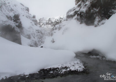 A snow-covered landscape features towering, jagged cliffs on either side, partially hidden by a blanket of mist. In the foreground, a smooth, white layer of snow undulates gently, contrasting with dark rocky formations peeking through. A narrow, winding stream can be seen at the bottom, with clear water flowing, creating a stark contrast against the surrounding snow. A solitary figure in dark clothing stands near the edge of the snow, adding a sense of scale to the vast, icy environment. The scene conveys a sense of tranquility and isolation in a winter wonderland.