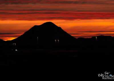 The image captures a silhouette of a mountain against a vibrant sunset. The sky is painted in rich hues of orange, red, and subtle hints of purple, creating a dramatic backdrop. There are also a few streetlights visible, their soft glow contrasting against the dark foreground and the outlines of the distant hills. The overall scene evokes a sense of calm and beauty in nature, emphasizing the grandeur of the landscape at dusk.