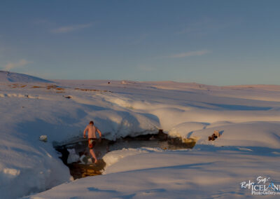 A person in swim trunks is walking towards a small, warm water spring surrounded by snow in a winter landscape. The ground is covered in thick, white snow, and the sky is clear with a hint of blue. There are some items laid out on the snow nearby, suggesting people are enjoying the natural hot spring. The scene captures the contrast between the cold environment and the warm spring.