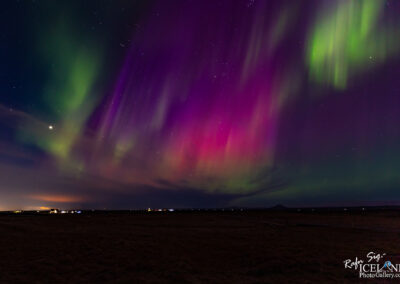 A stunning night sky illuminated by the aurora borealis, featuring vibrant colors of purple and green dancing across the dark canvas of the heavens. The atmospheric lights appear to shimmer and flow, creating a mesmerizing display. Below the vibrant auroras, a dark landscape is visible, with small lights from distant settlements twinkling in the background, adding a sense of perspective to the scenic beauty. The scene captures the magic of nature in a remote area, with stars scattered throughout the sky, enhancing the overall enchanting atmosphere.