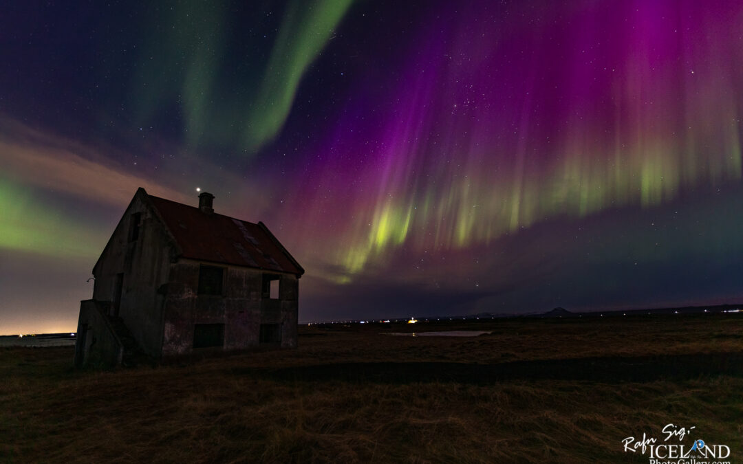 A solitary, weathered house with a slanted red roof stands in a vast, open landscape at night. The house has several windows, some of which appear dark and empty. In the background, the sky is illuminated by vibrant colors from the Northern Lights, showcasing swirling shades of green, purple, and hints of blue. The ground is covered with golden-brown grass, and distant lights can be seen twinkling along the horizon. The scene conveys a sense of tranquility and wonder, merging the mystery of the universe with the quiet isolation of the house.