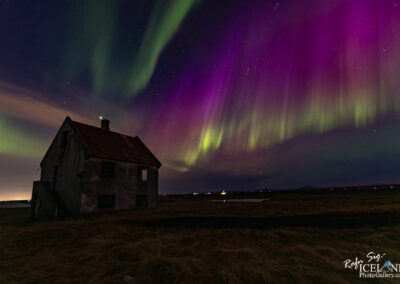 A solitary, weathered house with a slanted red roof stands in a vast, open landscape at night. The house has several windows, some of which appear dark and empty. In the background, the sky is illuminated by vibrant colors from the Northern Lights, showcasing swirling shades of green, purple, and hints of blue. The ground is covered with golden-brown grass, and distant lights can be seen twinkling along the horizon. The scene conveys a sense of tranquility and wonder, merging the mystery of the universe with the quiet isolation of the house.