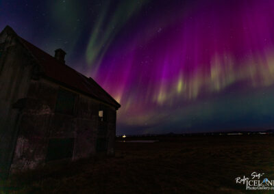 In this image, a rustic, abandoned building is positioned in the foreground, tilted slightly to one side. The structure has a weathered look, with dark walls and a rusty red roof. Above it, a stunning display of the Northern Lights illuminates the sky with vibrant colors, including deep purples, greens, and hints of blue. The auroras create flowing patterns that stretch across the night sky, dotted with twinkling stars. In the background, a soft, glowing light hints at distant areas, while the land below is flat and dark, enhancing the ethereal quality of the scene.
