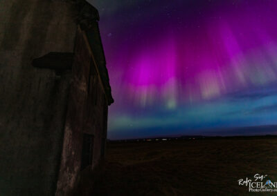 The image depicts a striking night sky illuminated by vibrant colors of the aurora borealis, featuring shades of purple, green, and blue. To the left, the crumbling wall of an old, weathered building is silhouetted against the colorful sky, creating a stark contrast. The background shows distant hills and a hint of lights from faraway structures, while the upper part of the image is filled with stars twinkling among the swirling aurora. The scene evokes a sense of tranquility and wonder, capturing the beauty of a natural light display in a remote landscape.