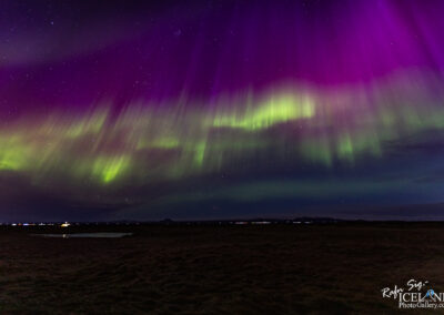 The image features a stunning display of the aurora borealis, with vibrant shades of purple and green swirling against a dark night sky. The auroral lights appear to ripple and flow, creating a dynamic, almost ethereal atmosphere. In the background, faint stars are visible, adding to the cosmic feel of the scene. The landscape below includes a grassy area, with a small reflective body of water, hinting at the tranquility of the surrounding environment. The horizon reveals distant lights, suggesting human presence far away.