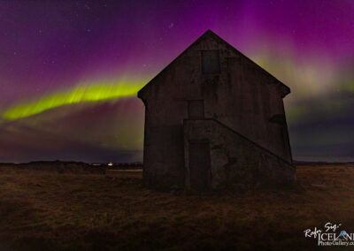 A dark, weathered house stands silhouetted against a vibrant night sky. The sky is illuminated with swirling shades of green and purple, suggesting the presence of the Northern Lights. Stars are scattered throughout the background, enhancing the celestial atmosphere. In the foreground, the grassy landscape is dimly visible, providing a sense of depth to the scene. The overall mood is tranquil yet awe-inspiring, capturing the beauty of nature’s light display.