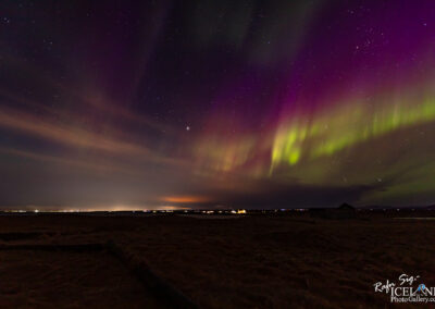 A stunning night sky filled with vibrant colors of purple and green dancing across the backdrop of stars, known as the Northern Lights. The ground below is dark and flat, with hints of a field and possibly some distant structures or lights in the background. The overall scene conveys a sense of tranquility and natural beauty, as the auroras create a mesmerizing display over an expansive landscape at night.