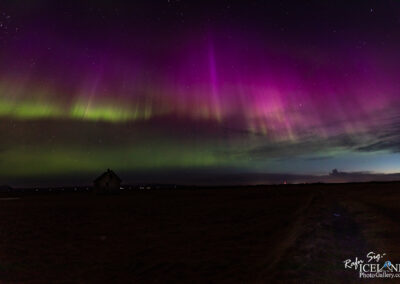 The image captures a stunning display of the northern lights, showcasing vibrant hues of purple and green swirling across the night sky. Below, a solitary, old farmhouse is silhouetted against the illuminated sky, adding a sense of scale and tranquility to the scene. The landscape is dark and expansive, with faint outlines of hills in the distance. Stars twinkle above, enhancing the serene and magical atmosphere created by the aurora borealis.