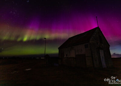 A silhouette of an old, rustic building with a slanted roof stands in the foreground, while the night sky above bursts with vibrant auroras. The display features swirling hues of purple and green, contrasted against a starry background. To the left, a lone light pole stands, casting a faint glow. The landscape is largely dark, suggesting a remote, tranquil setting where the natural spectacle of the auroras takes center stage.