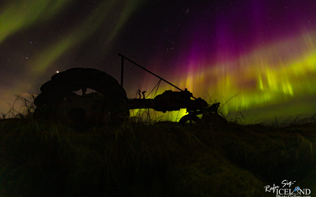 A silhouette of an old tractor sits in the foreground, partially obscured by tall grass. In the background, vibrant waves of green and purple light dance across the night sky, created by the aurora borealis. Stars are scattered throughout the dark sky, providing a sense of depth and vastness to the scene. The mood is serene and enchanting, as the natural light display contrasts beautifully with the rustic machinery.
