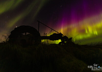 A silhouette of an old tractor sits in the foreground, partially obscured by tall grass. In the background, vibrant waves of green and purple light dance across the night sky, created by the aurora borealis. Stars are scattered throughout the dark sky, providing a sense of depth and vastness to the scene. The mood is serene and enchanting, as the natural light display contrasts beautifully with the rustic machinery.