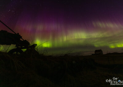 A stunning night sky filled with vibrant colors creates a breathtaking display. A rich palette of deep purples and vivid greens glows overhead, resembling curtains of light dancing in the darkness. The stars twinkle against the backdrop, enhancing the magic of the scene. In the foreground, a silhouette of a vintage bike rests on the ground, with patches of grass and an old, simple house in the distance. The entire composition conveys a serene and otherworldly atmosphere, inviting viewers to immerse themselves in the beauty of nature.