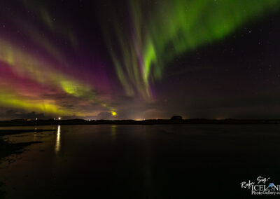The image captures a breathtaking display of the Northern Lights, with vibrant swirls of green, purple, and hints of yellow illuminating the night sky. Wispy tendrils of light dance across the horizon, creating a magical atmosphere against a backdrop of dark clouds and twinkling stars. Below, a calm body of water reflects the colorful lights above, adding depth to the scene. In the foreground, faint silhouettes of distant land and a small building can be seen, enhancing the sense of tranquility and natural beauty in this stunning nightscape.