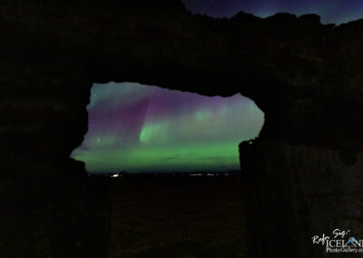 The image shows a vibrant display of the northern lights, characterized by swirling bands of green and purple hues lighting up the night sky. This beautiful phenomenon is framed by an irregular stone arch, creating a natural window to the scene. In the distance, there are faint lights from a building and other structures, contrasting with the dark landscape below. The sky is dotted with stars, adding to the serene atmosphere of the nighttime setting.