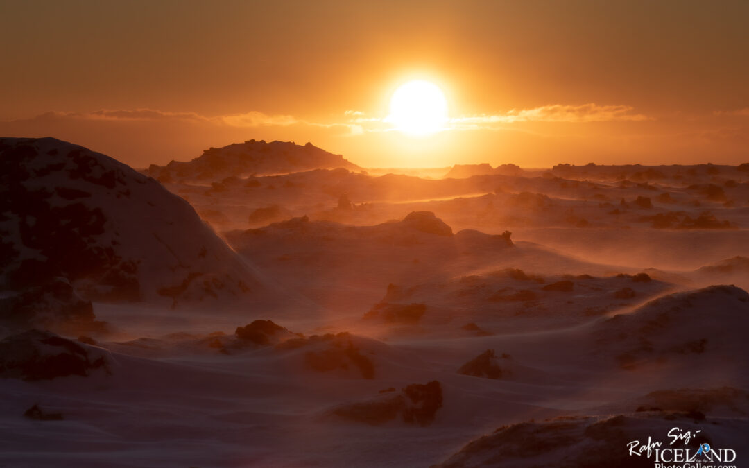 The image depicts a breathtaking sunset scene over a snow-covered landscape. The sun, large and golden, is setting on the horizon, casting warm orange and yellow hues across the sky. In the foreground, there are dark, rocky formations partially covered in snow, standing out against the glowing backdrop. The scene evokes a sense of chilly tranquility, with gentle snowdrifts and a hint of wind creating a soft, flowing effect in the surrounding snow. The overall atmosphere is peaceful and serene, capturing the beauty of nature at twilight.