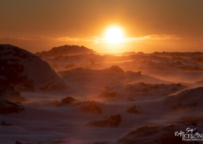 The image depicts a breathtaking sunset scene over a snow-covered landscape. The sun, large and golden, is setting on the horizon, casting warm orange and yellow hues across the sky. In the foreground, there are dark, rocky formations partially covered in snow, standing out against the glowing backdrop. The scene evokes a sense of chilly tranquility, with gentle snowdrifts and a hint of wind creating a soft, flowing effect in the surrounding snow. The overall atmosphere is peaceful and serene, capturing the beauty of nature at twilight.