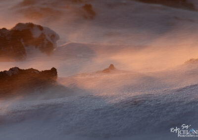 The image captures a dramatic landscape where wind-blown snow swirls around dark, rugged rocks. The scene is illuminated by soft, warm hues, suggesting a sunset or sunrise. Wisps of snow are caught in the air, creating a sense of movement and energy. The textures of the rocks contrast starkly with the smooth, flowing snow, while shadows add depth to the scene. Overall, it evokes a feeling of tranquility amidst the wildness of nature