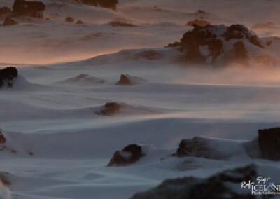 A serene winter landscape features gentle slopes covered in a thick blanket of soft, white snow. Dark, rugged rocks emerge sporadically from the snow, their textures contrasting sharply against the white surface. The scene is infused with a warm glow from a low-hanging sun, creating soft shadows and highlighting the contours of the snowdrifts and rocks. Wisps of wind swirl around, causing a light flurry of snow particles to dance in the air, adding a sense of movement to the peaceful ambiance. The overall atmosphere is quiet and calm, evoking a sense of untouched natural beauty.