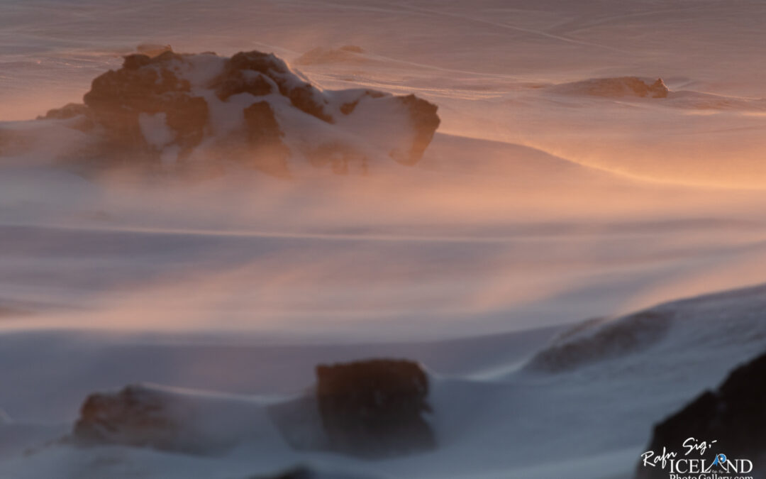 A vast and tranquil snowy landscape with soft hues of pink and orange illuminating the scene, likely at dawn or dusk. Gentle winds create patterns in the snow, while distant rocky outcrops emerge from the blanket of white. The atmosphere feels serene, with a sense of quiet beauty and the chill of cold air. The interplay of light and shadow enhances the rugged textures of the rocks, adding depth to the tranquil setting.