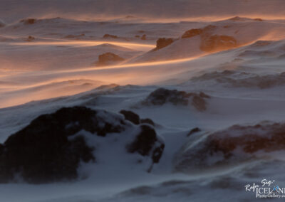 A winter landscape featuring gently rolling hills covered in snow, with stones peeking through. The scene captures a sense of movement, as light snow is being blown across the surface, creating soft, flowing lines in the white blanket. The horizon is lit with a warm, golden glow, suggesting the presence of a low sun. The overall atmosphere feels cold and serene, evoking the beauty and stillness of a wintry day.