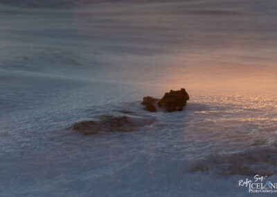 The image captures a tranquil scene of a shoreline at sunset. The foreground features several small, rugged rocks partially covered by gently lapping waves. The surface of the water appears frothy, with white foam contrasting against the dark shapes of the rocks. In the background, the soft light of the setting sun casts a warm, golden hue across the water, creating a serene and calming atmosphere. The gentle movement of the waves enhances the peacefulness of the scene.
