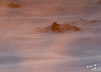 The image captures a landscape at dawn where waves of water gently lap against rocky formations. The scene is enveloped in soft, warm tones of orange and pink, creating a serene and dreamlike ambiance. The rocks, partially submerged or jutting out of the water, provide a rugged contrast to the smoothness of the water surface. A sense of tranquility permeates the image, evoking the sounds of gentle waves and a light breeze, making it an inviting representation of a peaceful coastal moment.