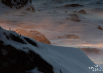 A serene winter landscape is depicted, featuring soft, swirling snow atop rocky terrain. The scene captures a gentle light, possibly from the setting sun, casting a warm glow on a circular rock in the center, contrasting with the cool icy tones surrounding it. Snowflakes can be seen caught in the breeze, creating a subtle sparkle in the air. The background fades into a misty distance, enhancing the tranquil ambiance of the snowy wilderness.