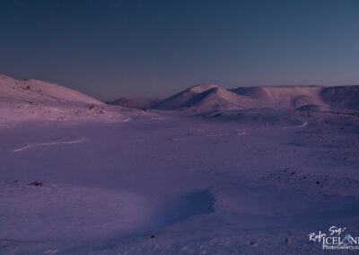 A vast, snow-covered landscape stretches out beneath a twilight sky. The ground is mostly smooth with subtle undulations, and patches of snow reflect a soft, muted purple hue. In the background, gentle hills rise and fall, their slopes blanketed in white. The horizon is faintly visible, blending into the darkening sky where deep blues transition to lighter shades near the horizon. The scene evokes a sense of tranquility and isolation, typical of a cold, quiet winter evening in a remote area.