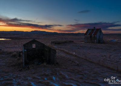 A desolate landscape at dusk featuring two dilapidated structures, one with a rusted roof, surrounded by dry grass and sparse vegetation. The sky is painted with shades of orange and blue as the sun sets, casting a tranquil glow over the scene. In the background, rolling hills are silhouetted against the colorful horizon, with a small body of water reflecting the sunset.