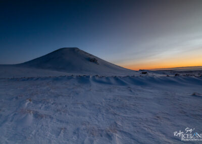 The image features a large, snow-covered hill or volcano that rises prominently from a flat, expansive landscape. The hill has a gentle slope leading to a smooth peak, while the surrounding area is blanketed in fresh, white snow. In the background, the sky transitions from a deep blue to lighter shades near the horizon, indicating either dawn or dusk. Soft lines of snow drifts create a textured foreground, and a few scattered rocks are visible in the snow. The overall atmosphere suggests a serene, winter setting in a remote location.
