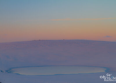 A serene winter landscape featuring a partially frozen lake surrounded by snow-covered terrain. The sky transitions from soft pink to pale blue, indicating dawn or dusk. Sparse hills are visible in the background, with a few dark silhouettes on the horizon. The scene evokes a sense of tranquility and the beauty of nature in a cold environment.