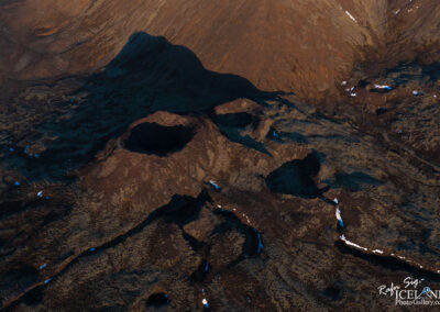 An aerial view of a volcanic landscape featuring several craters and rugged terrain. The ground is predominantly dark brown with patches of mossy green, and there are remnants of snow scattered throughout. The largest crater is near the center, showcasing a deep, circular shape, while smaller craters are visible around it. The terrain appears uneven with subtle ridges and shallow valleys, creating a dramatic and barren aesthetic typical of volcanic regions. The lighting enhances the texture of the land, emphasizing the stark contrast between the dark earth and the brighter snow patches.