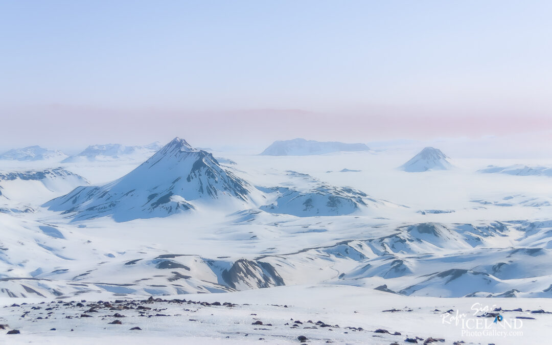 A vast, snow-covered landscape stretches out beneath a pale blue sky. In the foreground, the ground is blanketed in smooth, white snow with a few scattered dark rocks protruding. Beyond this, multiple mountains rise sharply, their peaks capped with snow and glistening in the sunlight. The mountains vary in shape and size, some steep and jagged while others are more rounded. The background features further distant peaks that fade into a soft mist, enhancing a sense of depth and solitude in this serene, wintry scene. The overall atmosphere suggests a peaceful yet majestic wilderness.