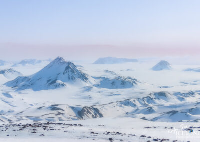 A vast, snow-covered landscape stretches out beneath a pale blue sky. In the foreground, the ground is blanketed in smooth, white snow with a few scattered dark rocks protruding. Beyond this, multiple mountains rise sharply, their peaks capped with snow and glistening in the sunlight. The mountains vary in shape and size, some steep and jagged while others are more rounded. The background features further distant peaks that fade into a soft mist, enhancing a sense of depth and solitude in this serene, wintry scene. The overall atmosphere suggests a peaceful yet majestic wilderness.